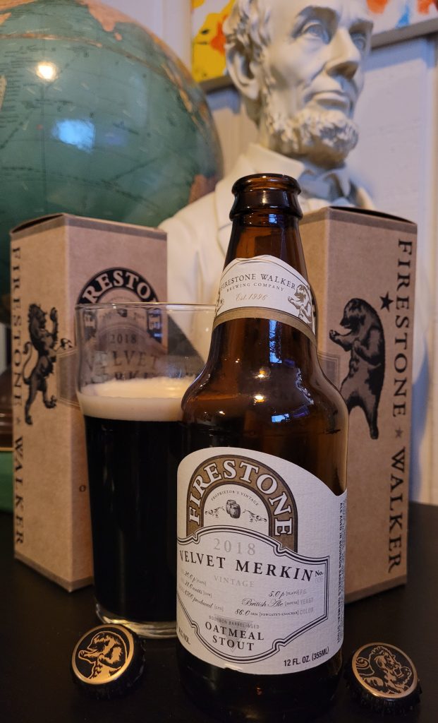 A bottle and full glass of the 2018 Velvet Merkin Barrel-Aged Oatmeal Stout by Firestone Walker. The bottle is flanked by the bottle's cardboard packaging and its bottle caps.