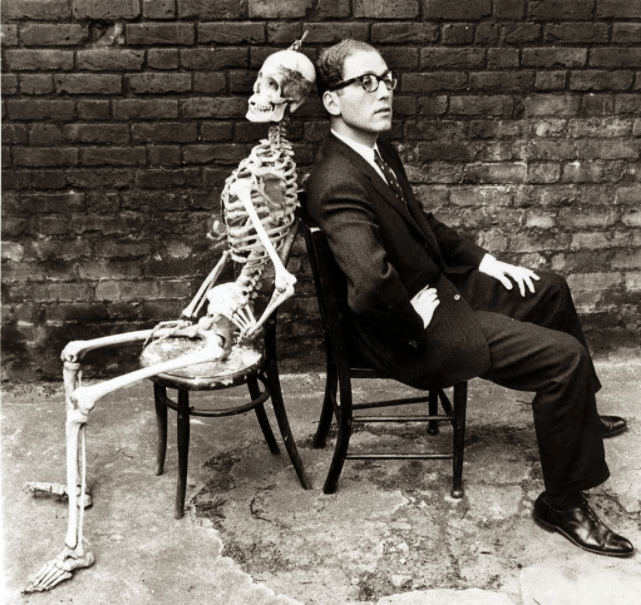 Black and white photo of Tom Lehrer sitting in a chair, back-to-back with an anatomical skeleton. 