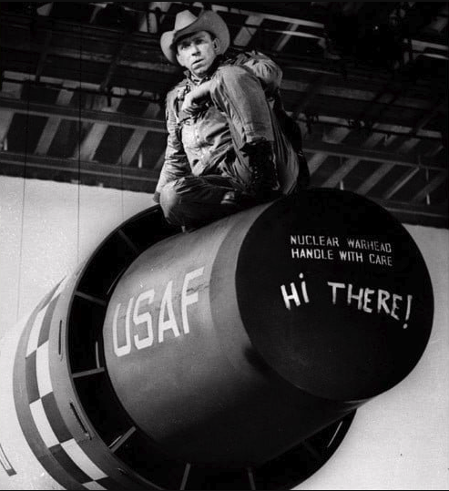 Black and white image of actor Slim Pickens as Major Kong sitting atop a prop version of a USAF nuclear warhead. Painted on the warhead are the words Nuclear Warhead Handle With Care. Below that, someone else has painted the words Hi There!