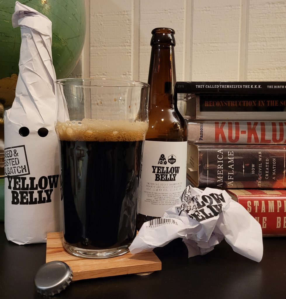 Image of two bottles of Yellow Belly Imperial Stout -- the one on the left remains in its white paper wrapper, the one on the right is unwrapped and opened. Between the two bottles is a beer glass three-fourths full with a nearly-black beer that has a dark tan head. Behind the opened bottle on the right is a stack of books referenced in this episode.