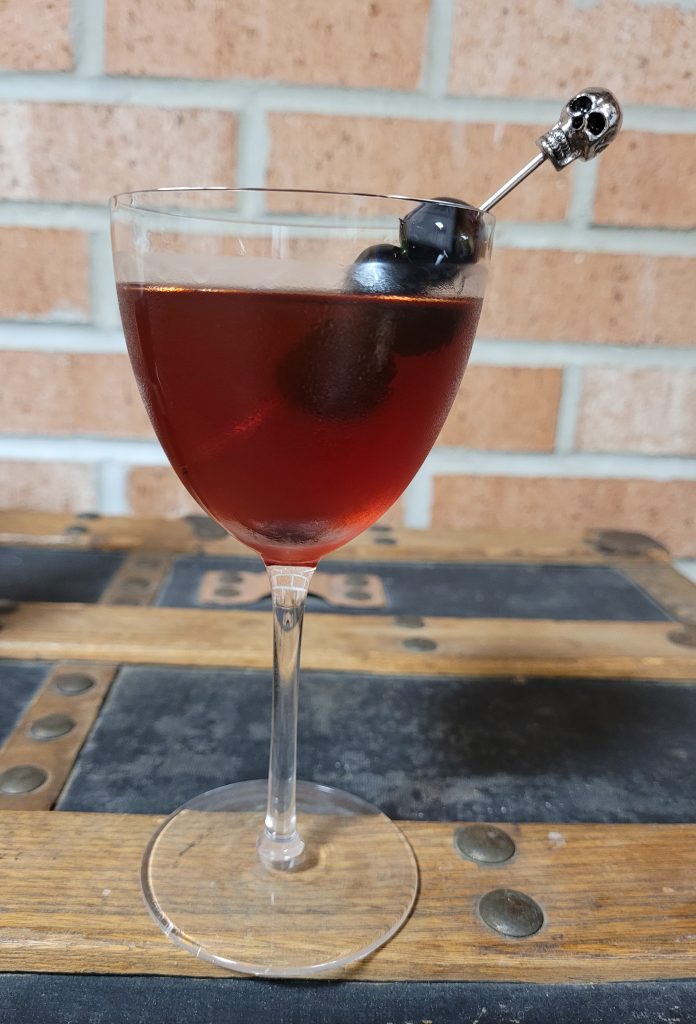 Photo of the mystery drink in a Nick and Nora glass with three Luxardo Maraschino cherries on a chrome cocktail pick topped by a tiny chrome skull. 