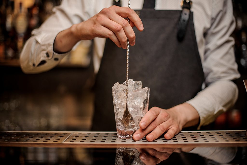 Photo of a bartender's hands holding a glass full of ice cubes in one hand and a cocktail spoon in the other and is read to stir. 