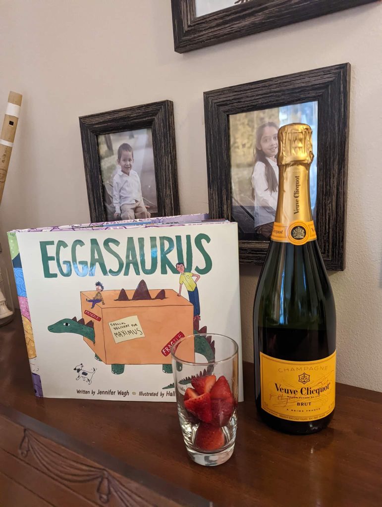 Image of a bottle of Veuve Clicquot, a glass of cut strawberries, and a copy of Eggasaurus